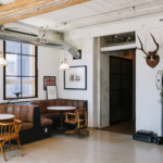 A modern industrial-style room with a seating area featuring a brown leather booth, a wooden chair, and round tables. Exposed pipes, wall art, a deer head mount, and pendant lights enhance the space's rustic charm. A window provides natural light.