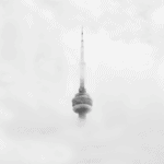 A tall, needle-like tower emerges through thick fog, with its bottom partially obscured by the mist. The structure is stark against a white sky, creating a serene and mysterious atmosphere.