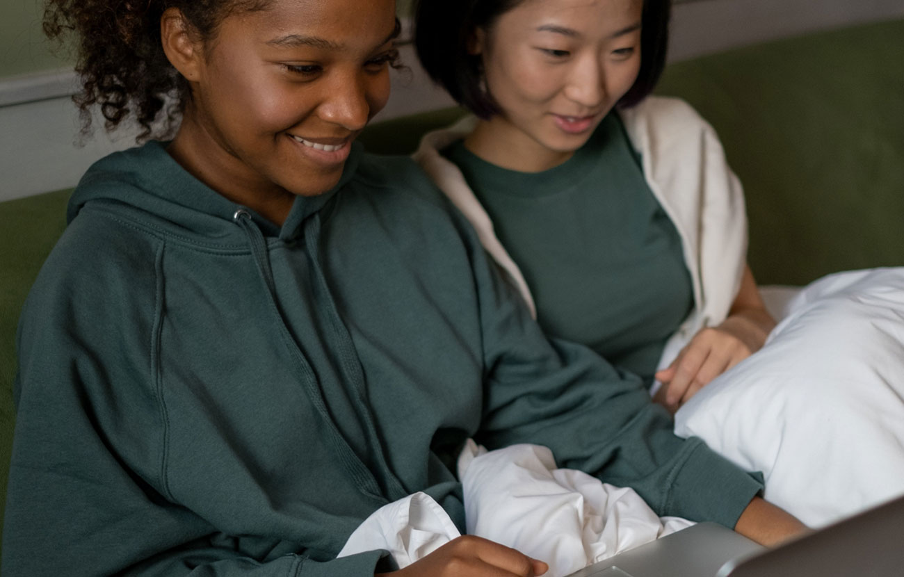 Two people sitting closely together on a couch, smiling while looking at an open laptop. They are wrapped in white blankets and wearing comfortable, casual clothing. The atmosphere appears cozy and relaxed.