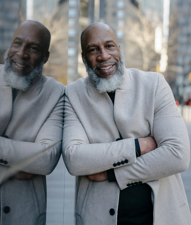 A man with a gray beard and wearing a light gray coat smiles while leaning against a reflective glass surface. Urban buildings and a leafless tree are visible in the background.