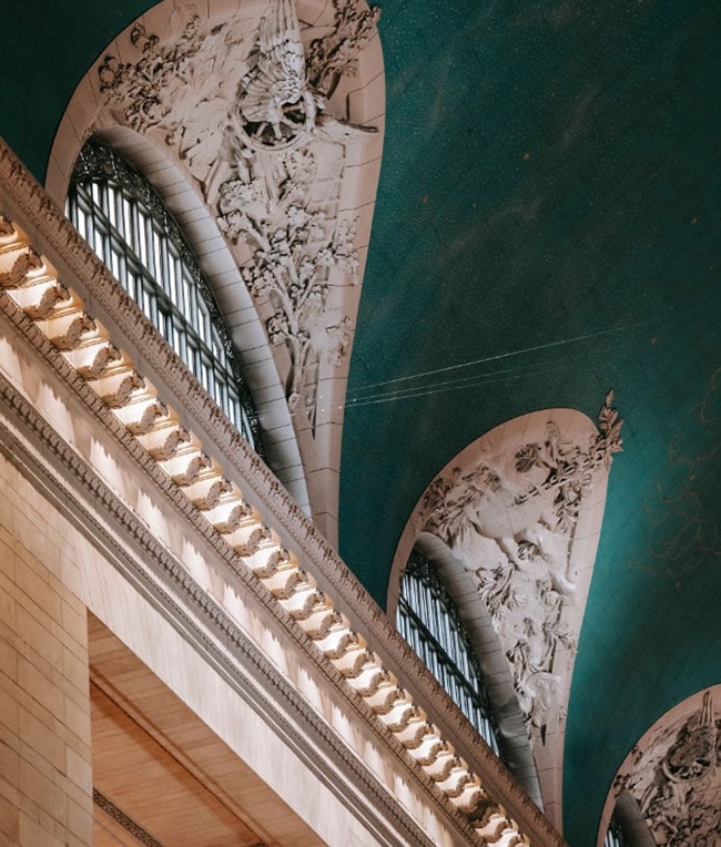 Close-up of the ornate ceiling and arched windows of a historic building. The ceiling features intricate relief sculptures and a teal background, bathed in natural light.