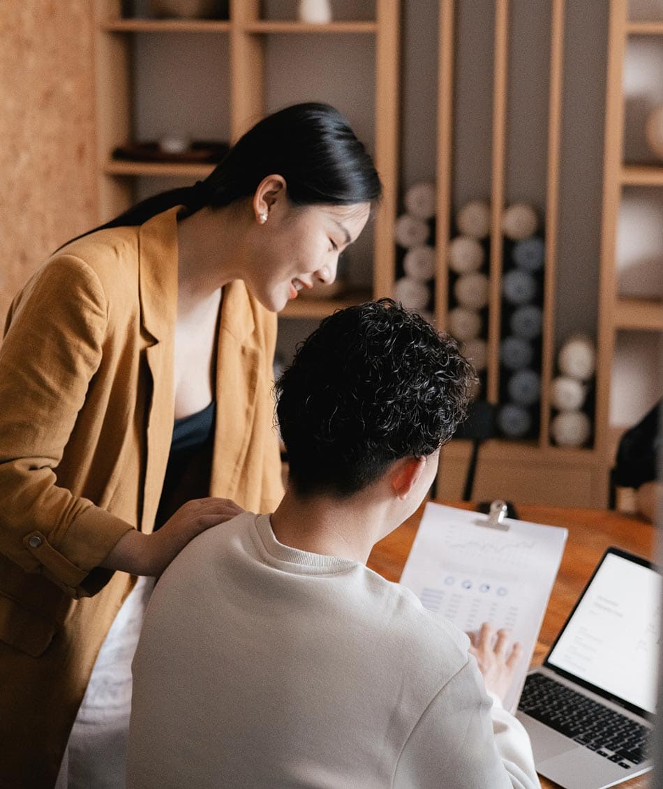 Two people collaborate in an office. One stands, leaning over to view a document held by the seated person, who is also using a laptop. Shelving with cylindrical objects is in the background, creating a cozy work atmosphere.