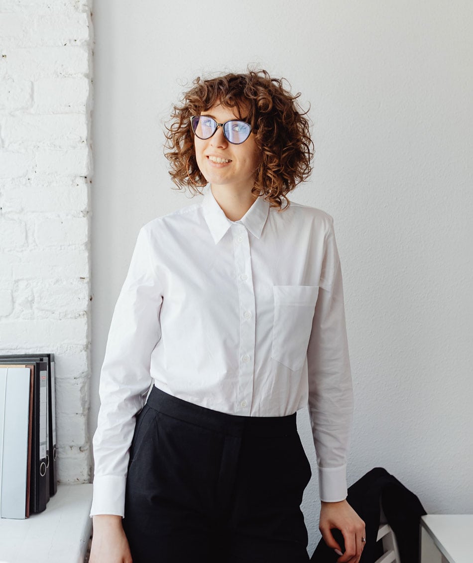 A person with curly hair and glasses stands smiling in an office, wearing a white shirt and black pants. They are leaning against a white brick wall, with binders and a desk visible nearby.