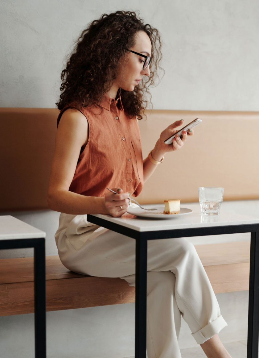 A woman with curly hair and glasses is sitting at a table, looking at her smartphone. She is wearing a sleeveless rust-colored top and light pants. There is a plate with a dessert and a glass of water on the table.