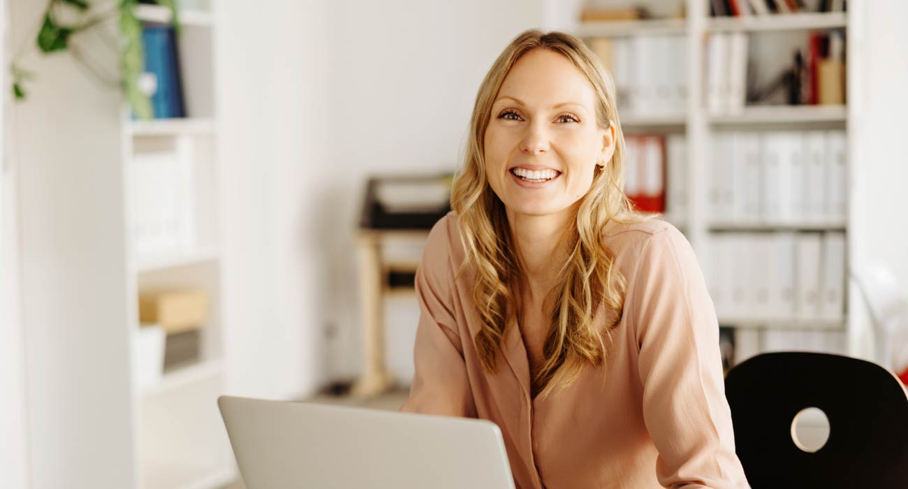 A smiling woman with long blonde hair sits at a desk in an office, working on a laptop. Bookshelves with various files and books are visible in the background.