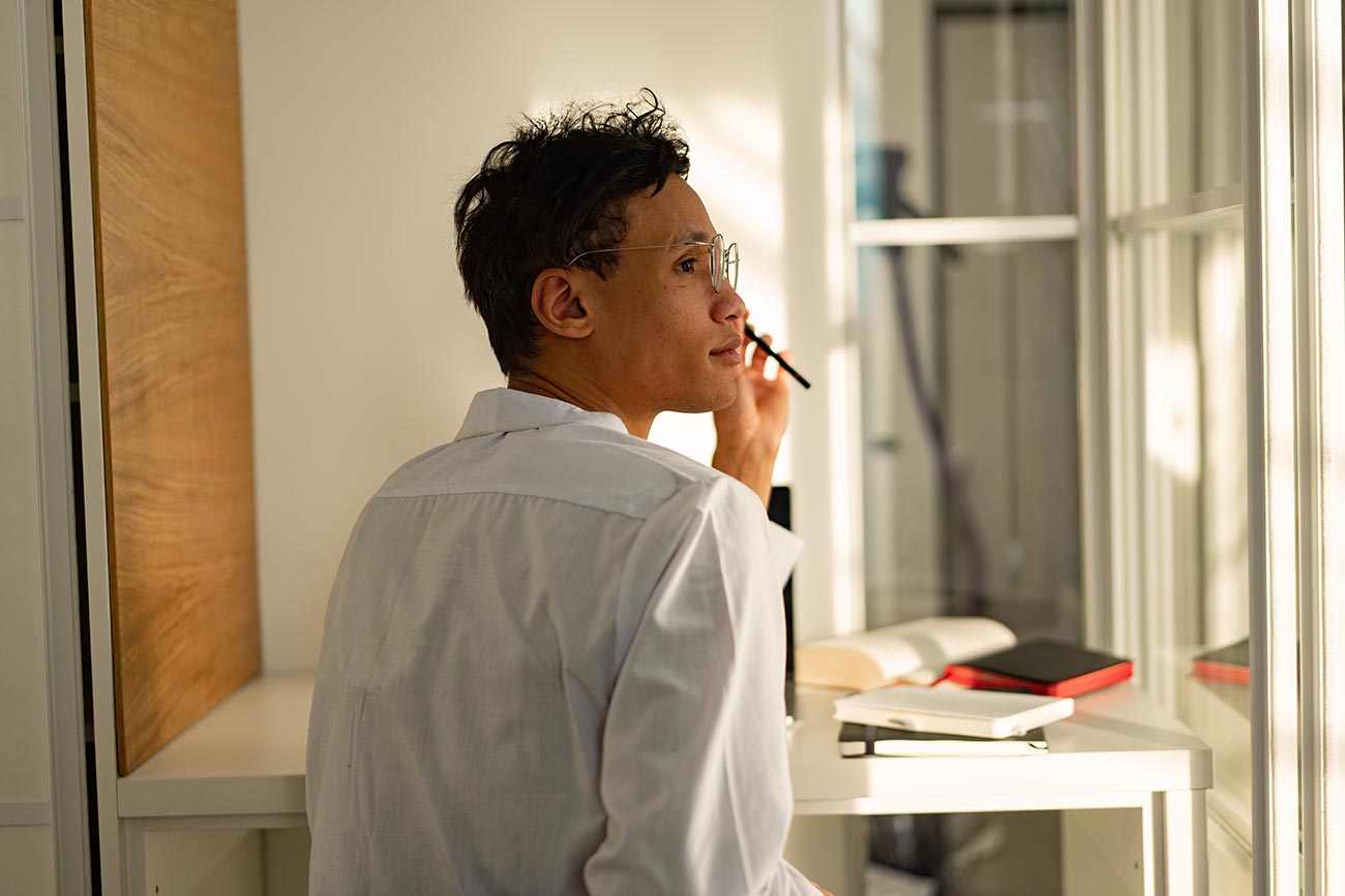 A person wearing a white shirt sits at a desk, looking thoughtfully out of a window. They hold a pen near their mouth, surrounded by open books and notebooks. Sunlight illuminates the room, casting soft shadows.