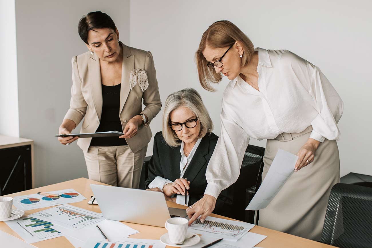 Three women in business attire work together in an office. One seated with a laptop, while the other two stand, examining documents. Charts and graphs are spread across the desk, and a cup of coffee is nearby. They appear focused and engaged in discussion.