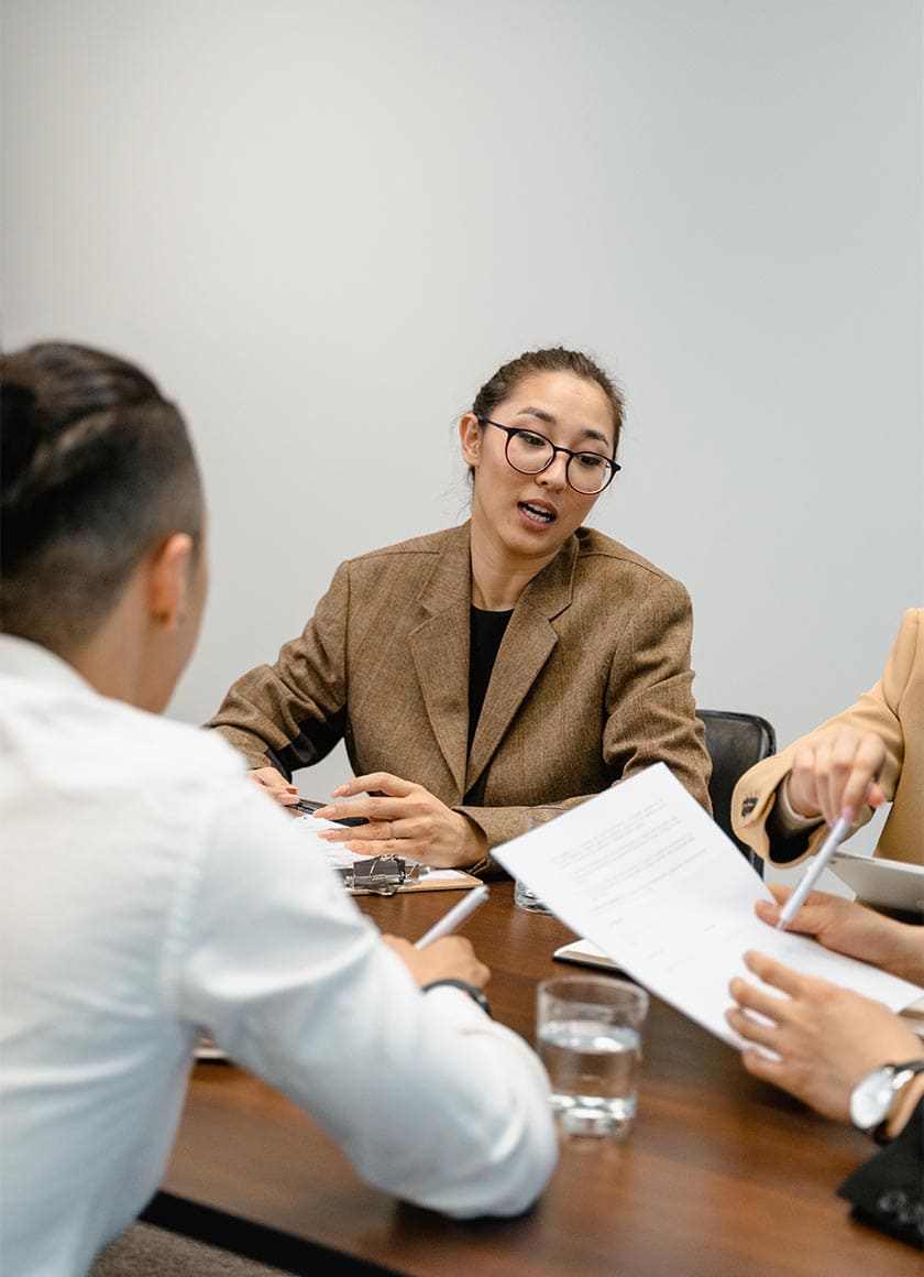 A person wearing glasses and a brown blazer is speaking while sitting at a table in a meeting. Two other individuals are partially visible, holding documents. Glasses of water are on the table. The atmosphere is professional.