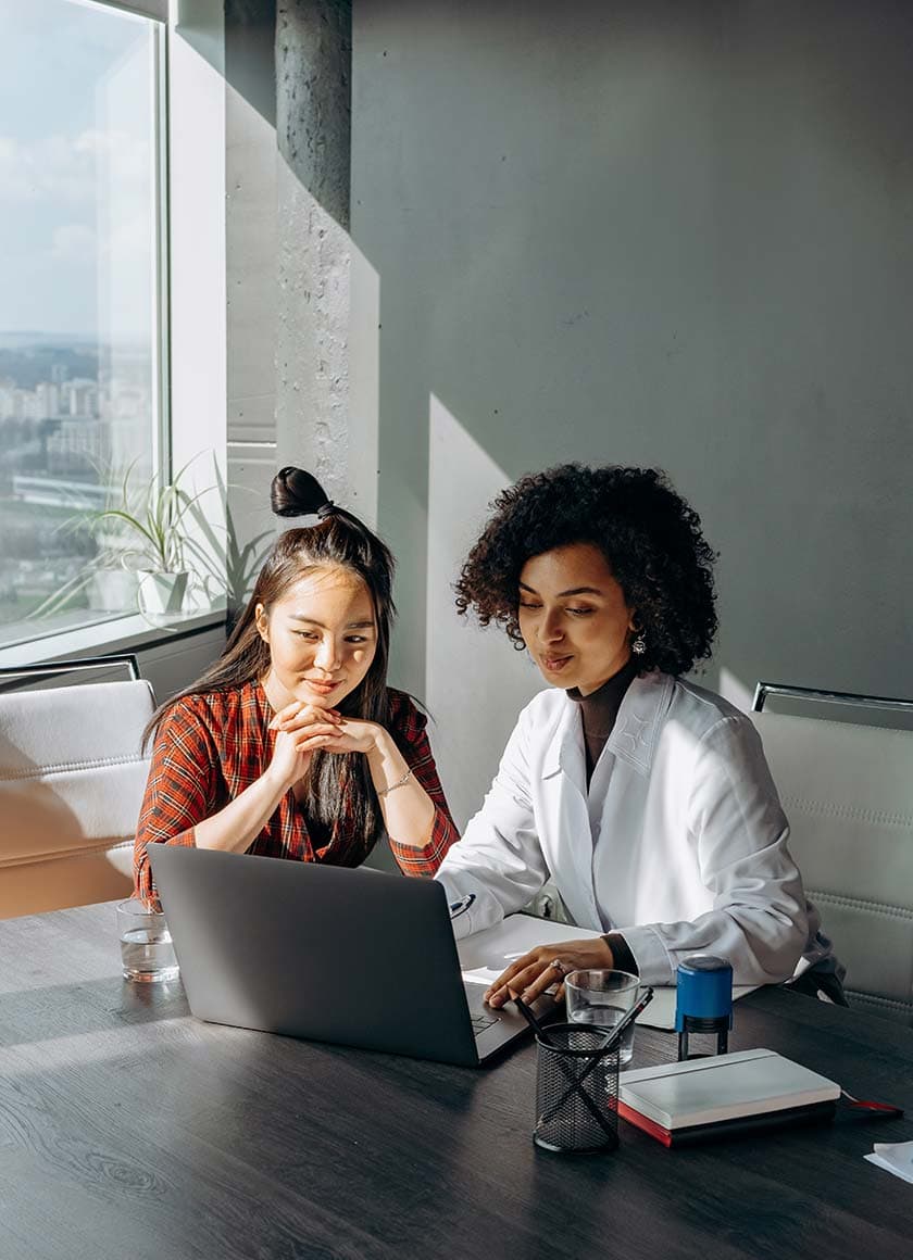 Two women sit at a table, looking at a laptop. One is in a white jacket, and the other wears a red plaid shirt. They appear focused and engaged in discussion. The room is well-lit by natural light from a large window.