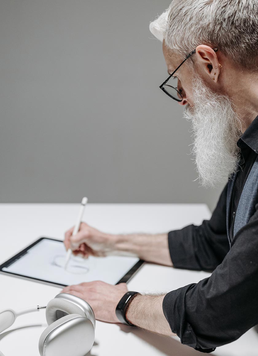 A person with a long white beard and glasses uses a stylus to draw on a tablet at a desk. A pair of wireless headphones rests on the table nearby.