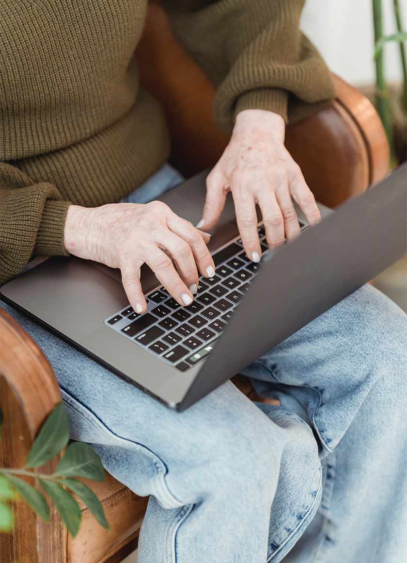 Person wearing a green sweater and blue jeans, sitting in a brown chair, typing on a laptop placed on their lap. The setting appears cozy, with plants visible in the foreground.