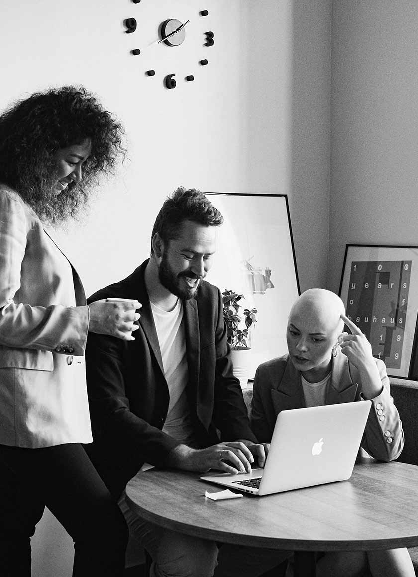 Three people are gathered around a laptop on a wooden table in a stylish room. One stands while the other two sit, engaged in discussion. A modern clock and framed art are visible on a nearby wall. The scene is black and white.