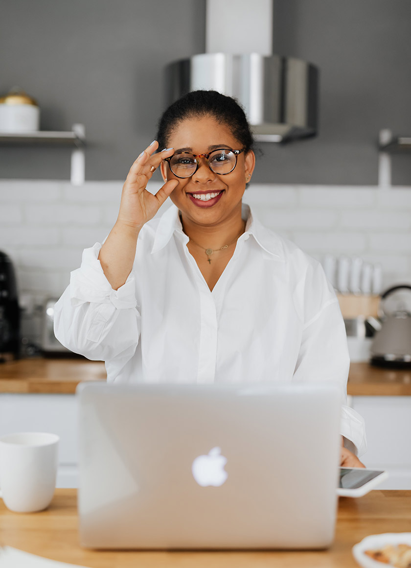 A person wearing glasses and a white shirt smiles while sitting at a kitchen table with an open laptop. They are holding a cup in one hand. The background features a white brick wall, wooden shelves, and kitchen items.